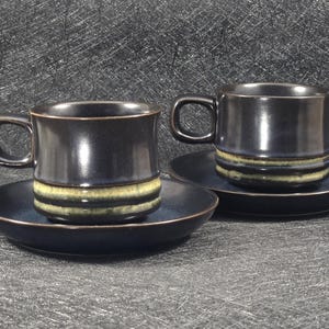 May include: Two dark blue ceramic coffee cups with a light brown stripe design, each sitting on a matching saucer.