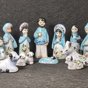 May include: A ceramic nativity scene with figures in blue and white robes with floral details. The set includes Mary, Joseph, baby Jesus, and other characters, plus animals such as sheep and a donkey. The figures are hand-painted.