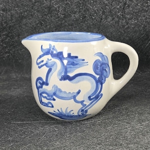 Peut inclure: Un pichet à crème en céramique de couleur crème orné d'un motif de cheval bleu peint à la main sur le côté. Le pichet présente un corps arrondi, un petit bec verseur, une anse incurvée et un liseré bleu sur le bord supérieur de l'ouverture.