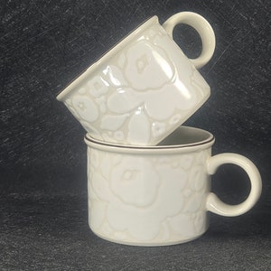 May include: Two white ceramic mugs with a floral design and brown trim. The mugs are stacked on top of each other.