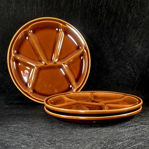 May include: Two brown ceramic divided serving plates. One plate is upright, showing five sections, while two more plates are stacked in the foreground. The plates have a glossy finish and a simple design, suitable for serving appetizers or snacks.