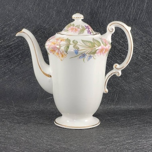 Peut inclure: Théière en céramique blanche avec un motif floral dans les tons de rose, jaune et bleu. La théière est ornée d'une bordure dorée autour du couvercle et de la base.