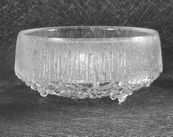 May include: A clear glass bowl with a textured, vertical pattern on the sides and a bubbly, rounded base. The bowl has a low profile and is likely used for serving or display. The glass appears to be thick and durable.