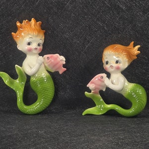 May include: Two ceramic mermaid figurines with pink fish in their hands. The mermaids have green tails and orange hair.