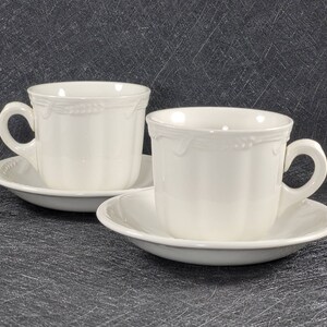 Peut inclure: Deux tasses à thé et soucoupes en céramique blanche avec un motif floral délicat. Les tasses ont une forme arrondie et une petite poignée. Les soucoupes sont légèrement plus grandes que les tasses et ont un bord festonné.