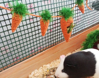 May include: Two guinea pigs in a cage with a green and orange crocheted carrot toy hanging from the top. One guinea pig is eating from a bowl of food, while the other is in a green and orange crocheted tunnel.