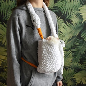 May include: A white crocheted swan-shaped pet carrier with an orange beak and a small white furry animal inside. The carrier is worn as a sling around a person's neck.
