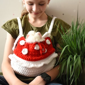 May include: A young person wearing a green shirt holds a crocheted red and white mushroom-shaped pet carrier with a white furry animal inside.