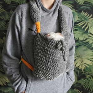 Peut inclure: Un porte-bébé en crochet gris avec un petit motif de canard orange et gris. Un petit rongeur blanc et gris est à l'intérieur du porte-bébé.