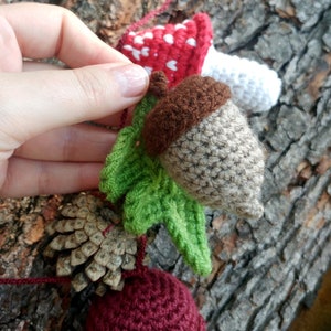 Peut inclure: Une guirlande en crochet avec un champignon rouge et blanc, un gland brun avec des feuilles vertes, une pomme de pin et un champignon bordeaux et blanc.