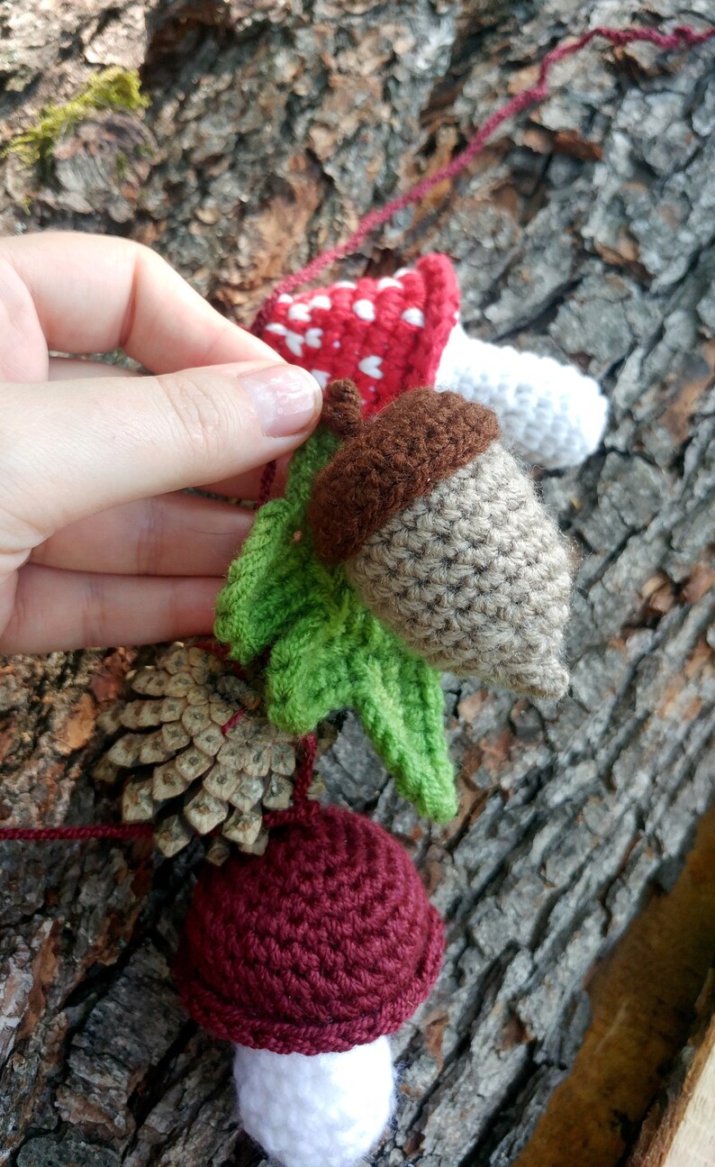 Peut inclure: Une guirlande en crochet avec un champignon rouge et blanc, un gland brun avec des feuilles vertes, une pomme de pin et un champignon bordeaux et blanc.