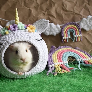 May include: A white crocheted unicorn-shaped pet bed with a small white guinea pig inside. The bed is decorated with colorful flowers and a horn. A rainbow-colored crocheted rainbow is in the foreground.