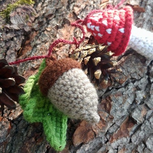 Peut inclure: Une guirlande de cr&eacute;atures de la for&ecirc;t en crochet, comprenant un champignon rouge et blanc, un gland brun et une pomme de pin, enfil&eacute;es sur un fil rouge.