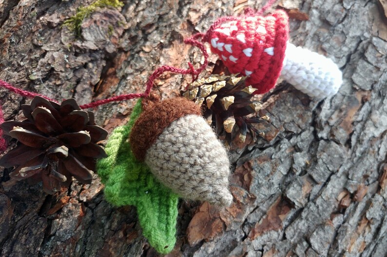 Peut inclure: Une guirlande de cr&eacute;atures de la for&ecirc;t en crochet, comprenant un champignon rouge et blanc, un gland brun et une pomme de pin, enfil&eacute;es sur un fil rouge.