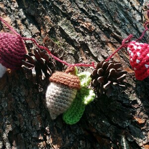 Peut inclure: Une guirlande en crochet avec des champignons rouges et blancs, des glands bruns et des pommes de pin. La guirlande est enfil&eacute;e sur un fil rouge et pend contre un tronc d'arbre.