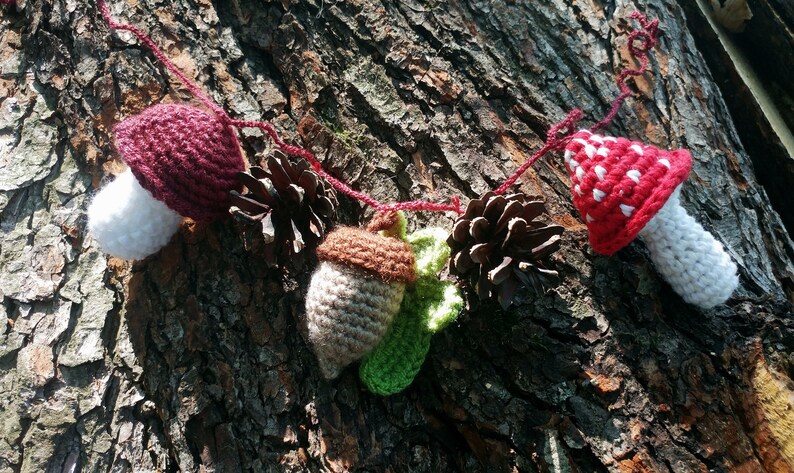 Peut inclure: Une guirlande en crochet avec des champignons rouges et blancs, des glands bruns et des pommes de pin. La guirlande est enfil&eacute;e sur un fil rouge et pend contre un tronc d'arbre.