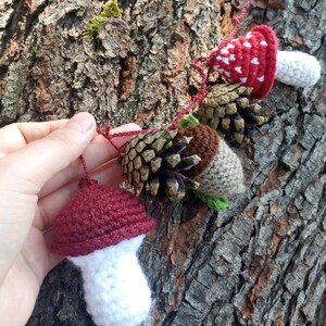 Peut inclure: Une guirlande en crochet avec des champignons rouges et blancs, des glands bruns et des pommes de pin. La guirlande est accroch&eacute;e &agrave; un tronc d'arbre.
