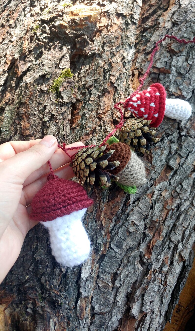 Peut inclure: Une guirlande en crochet avec des champignons rouges et blancs, des glands bruns et des pommes de pin. La guirlande est accroch&eacute;e &agrave; un tronc d'arbre.