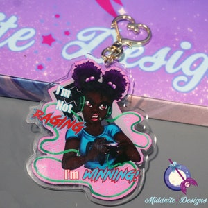Op de afbeelding: Een roze en groene acryl sleutelhanger met een cartoonillustratie van een zwarte vrouw met paars haar die een koptelefoon draagt en een videogamecontroller vasthoudt. De tekst "I'm Not Raging I'm Winning!" staat op de sleutelhanger gedrukt.