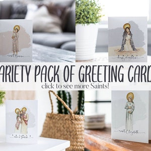 May include: Variety pack of greeting cards featuring watercolor illustrations of saints. The cards are white with a light blue background and feature the names of the saints in black text. The cards are displayed on a wooden table with plants and other decor.