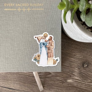 May include: A sticker featuring an illustration of two figures in religious attire, one holding a baby. The sticker is placed on a gray journal with the gold text "EVERY SACRED SUNDAY". Decorative yellow flowers are at the bottom of the sticker.