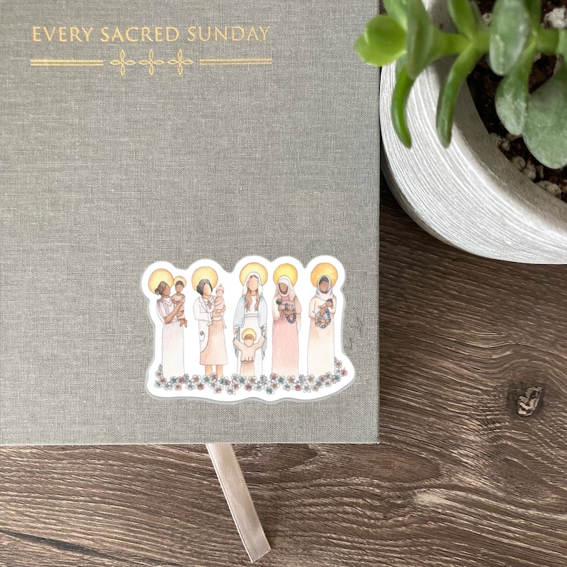 May include: A sticker featuring an illustration of several women holding babies, each with a halo. The sticker is placed on a gray journal with the gold text "EVERY SACRED SUNDAY". The sticker has a floral border.