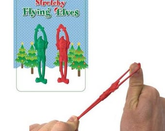 May include: A package of Stretchy Flying Elves toys. The package has a blue background with white polka dots and the words "Stretchy Flying Elves" in green and red. The package shows a green and a red elf. A red elf toy is being stretched.