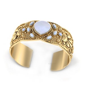 May include: A gold cuff bracelet with a large white mother-of-pearl gemstone and smaller white gemstones around it. The bracelet has a floral design.
