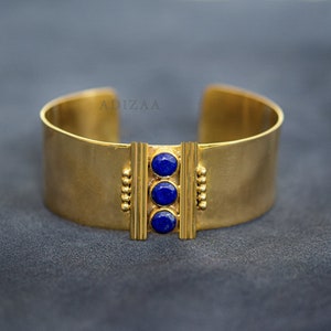 Può includere: Un braccialetto rigido in oro con tre accenti di pietre preziose blu. Il braccialetto è largo e ha un design semplice.