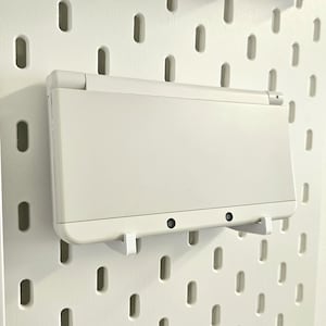 May include: A white handheld gaming console with a white pegboard background. The console is hanging on the pegboard with two white hooks.