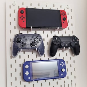 Peut inclure: Un panneau perforé blanc avec une Nintendo Switch Lite noire et bleue, une manette Playstation 4 noire et grise, une manette Nintendo Switch en plastique transparent et une console Nintendo Switch rouge et noire.