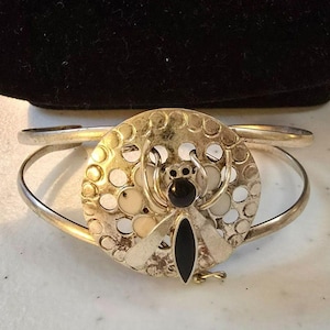 Puede incluir: Brazalete de puño plateado con un diseño de abeja circular. La abeja tiene un cuerpo y una cabeza negros, con detalles en blanco y crema. El brazalete tiene un diseño simple y elegante.