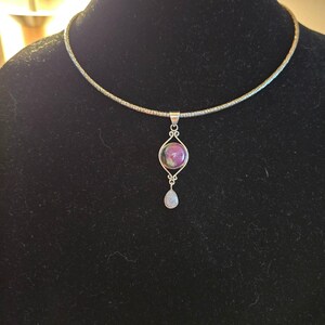 May include: A silver-toned necklace featuring a round, iridescent purple and green cabochon set within a decorative metal frame. A small, teardrop-shaped white stone hangs from the bottom of the frame, suspended by a textured silver-toned chain.