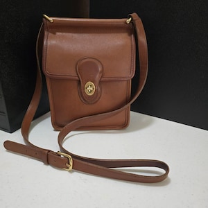 May include: A brown leather crossbody bag featuring a rectangular silhouette, a fold-over flap with a brass turn-lock closure, and an adjustable shoulder strap with a brass buckle. The bag has a smooth finish and visible stitching along the edges.