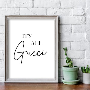 May include: A framed print with the text "It's All Gucci" in black script font on a white background. The frame is a light gray color and is sitting on a wooden surface.