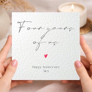 May include: A square white greeting card with a textured surface featuring the black cursive text Four years of us, a small red heart icon, and the printed text Happy Anniversary Alex in a simple black font centered at the bottom of the card.