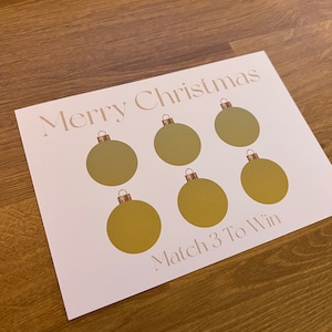 May include: A white card with the words "Merry Christmas" and "Match 3 To Win" in gold lettering. Six gold Christmas ornament shapes are printed on the card, suggesting a holiday game.