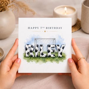 May include: A square greeting card with the text HAPPY 7TH BIRTHDAY in dark blue font. Below, the name RILEY is written in 3D letters patterned like footballs, positioned in front of a football goal on a green field with a light blue sky.
