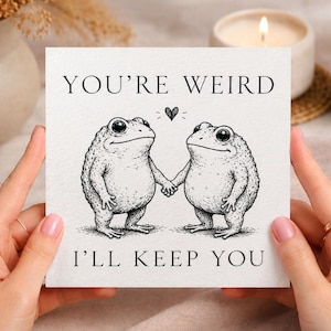 May include: A square greeting card with a white background featuring a black ink illustration of two frogs holding hands. Above the frogs is a small heart, and the text reads YOU'RE WEIRD at the top and I'LL KEEP YOU at the bottom in black font.