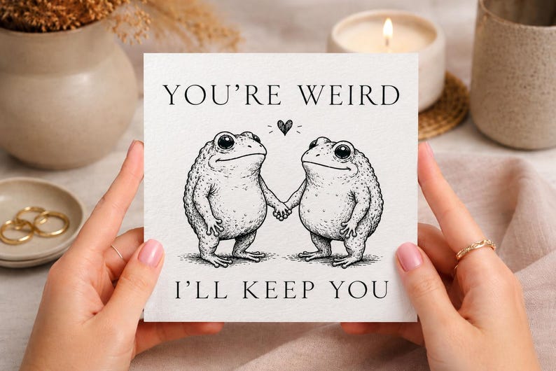 May include: A square greeting card with a white background featuring a black ink illustration of two frogs holding hands. Above the frogs is a small heart, and the text reads YOU'RE WEIRD at the top and I'LL KEEP YOU at the bottom in black font.