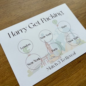 May include: A white card with the text "Harry Get Packing" printed in black. The card has six circles with the names of cities printed inside: London, New York, Paris, New York, Paris, and New York. The text "Match 3 To Reveal" is printed at the bottom of the card.