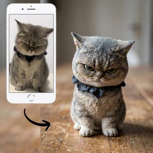 Puede incluir: Figura detallada de un gato gruñón con un collar negro. El gato tiene un patrón atigrado gris y marrón y está sentado sobre una superficie de madera. Un smartphone muestra una foto del mismo gato.