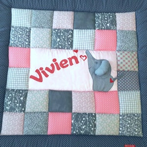 Puede incluir: Una colcha de patchwork cuadrada con un borde azul oscuro de lunares y un panel central rosa claro. El panel muestra el nombre Vivien en letras rojas de lunares, dos corazones rojos pequeños y una aplicación de elefante gris con un corazón.