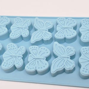 Puede incluir: Un molde de silicona azul claro con cavidades en forma de mariposa. El molde tiene forma rectangular con múltiples impresiones de mariposas. Cada mariposa tiene patrones de alas detallados, perfecto para manualidades o repostería.