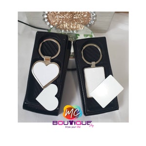 May include: Two keychains in black boxes. One is heart-shaped, the other rectangular, both with silver-tone metal frames and key rings. Each keychain has a white insert. A small white heart and rectangle are next to each keychain. The logo "MC Boutique" is at the bottom.