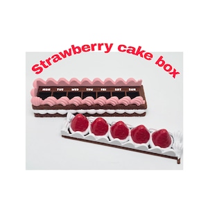Puede incluir: Una caja de pastel de fresa en miniatura con base de chocolate, glaseado rosa y una fila de fresas. La caja está etiquetada con los días de la semana. El texto "Strawberry cake box" es rosa.