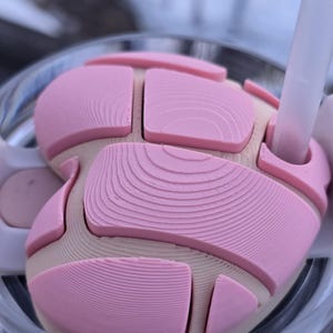 May include: Close-up of a pink and beige drink lid with a straw. The lid has a layered design with curved and rectangular sections. The straw is white. The lid appears to be 3D-printed and sits atop a clear glass.