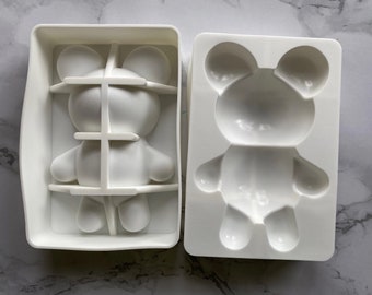 May include: Two white plastic molds for making bear-shaped ice cubes or treats. One mold is open, and the other is closed with a grid-like structure inside. The molds are designed with a bear's head, body, and limbs.