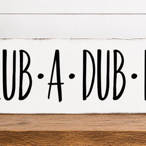 May include: A rectangular, distressed white wooden sign with the phrase "SCRUB • A • DUB • DUB" in black lettering. The sign is on a wooden surface, with a small potted plant and rolled white towels visible.