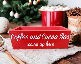 Könnte beinhalten: Rote Holzkiste mit dem Text "Coffee and Cocoa Bar warm up here" in weißen Buchstaben. Die Kiste steht auf einer weißen, flauschigen Oberfläche.